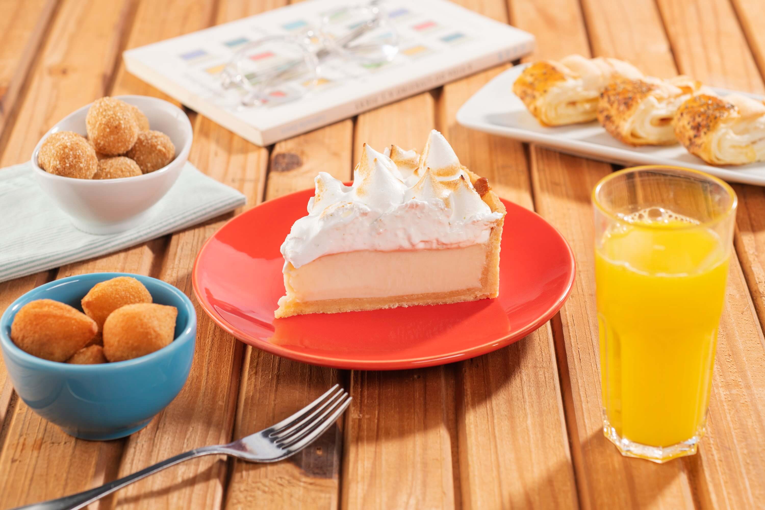 Bows com coxinhas, croquetes, folhados e um prato com torta de limão, ao lado de um copo com suco de laranja e um livro ao fundo.